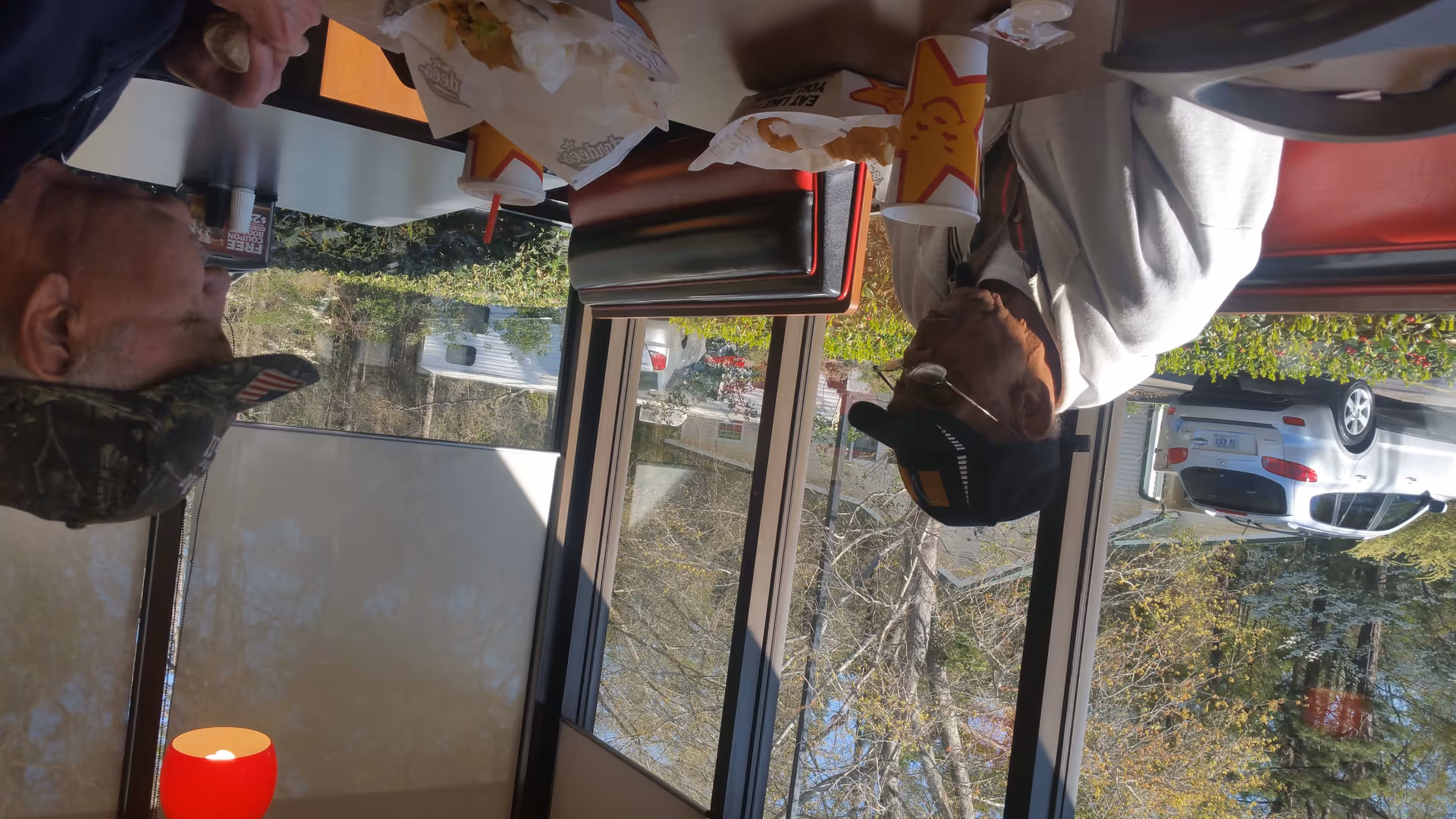 Two older men wearing caps sit at a restaurant booth by large windows eating fast food with cars visible outside.