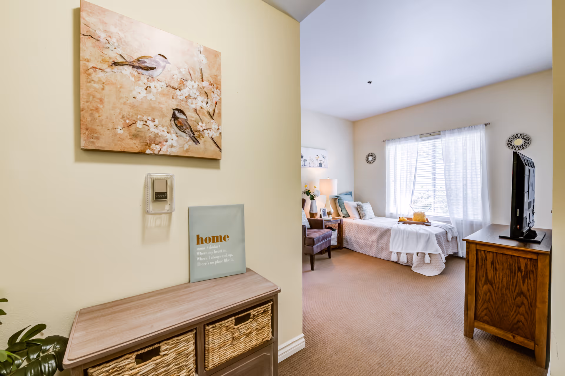 A cozy senior living bedroom with a single bed covered in light-colored bedding and decorative pillows. A wooden nightstand with a lamp and framed photo sits beside the bed. A wooden dresser with a flat-screen TV on top is positioned opposite the bed. The room has beige carpet and light-colored walls, with a large window covered by sheer white curtains allowing natural light to fill the space. Near the entrance, there is a small wooden cabinet with woven baskets and a decorative sign that reads 'home' along with a painting of birds on the wall above.