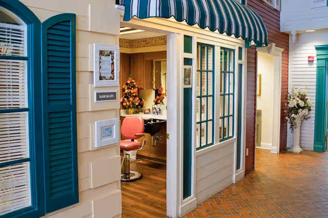 Interior view of a memory care facility hallway with a small hair salon featuring a red salon chair, a counter with a mirror, and floral decorations. The hallway has a brick-patterned floor and walls painted to resemble house fronts with windows and shutters.