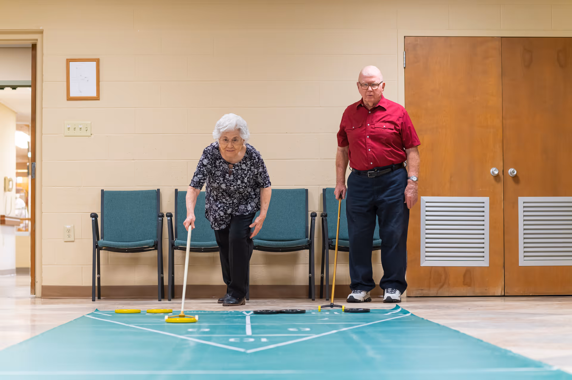 An elderly woman and man playing shuffleboard indoors in a senior living facility. The woman is actively sliding a shuffleboard puck while the man stands nearby holding a cue stick. Behind them are green chairs against a beige wall with wooden double doors to the right.