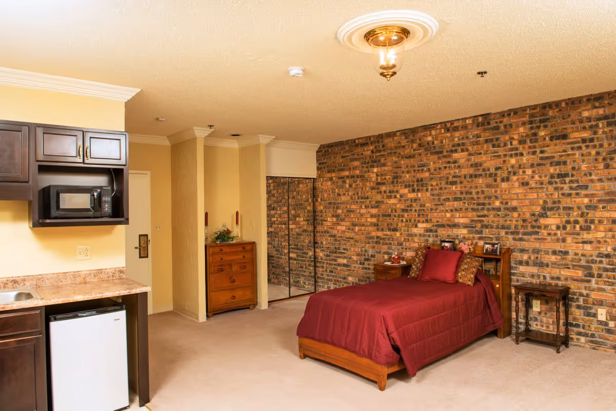 Interior view of a senior living facility room featuring a single bed with a red comforter and pillows against a brick accent wall. The room includes a wooden dresser, a small side table, and a kitchenette area with a microwave, mini fridge, and sink. There is a mirrored closet door and a ceiling light fixture.