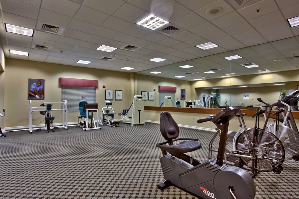 A fitness room with various exercise equipment including stationary bikes, weight machines, and other workout apparatus. The room has a patterned carpet, beige walls, a large mirror covering one wall, and fluorescent ceiling lights. There are framed pictures and a window with a pink valance on the far wall.