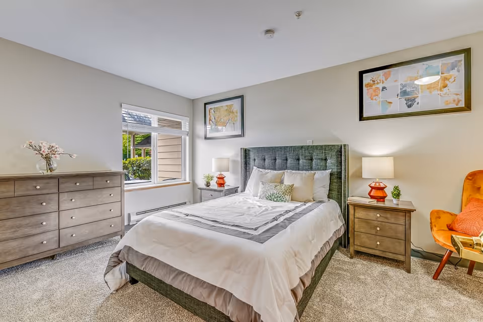 A bright and cozy bedroom featuring a neatly made bed with white and gray bedding, a tufted green headboard, two wooden nightstands with lamps, a wooden dresser with a vase of flowers, a large window letting in natural light, a colorful armchair with an orange pillow, and framed artwork on the walls.