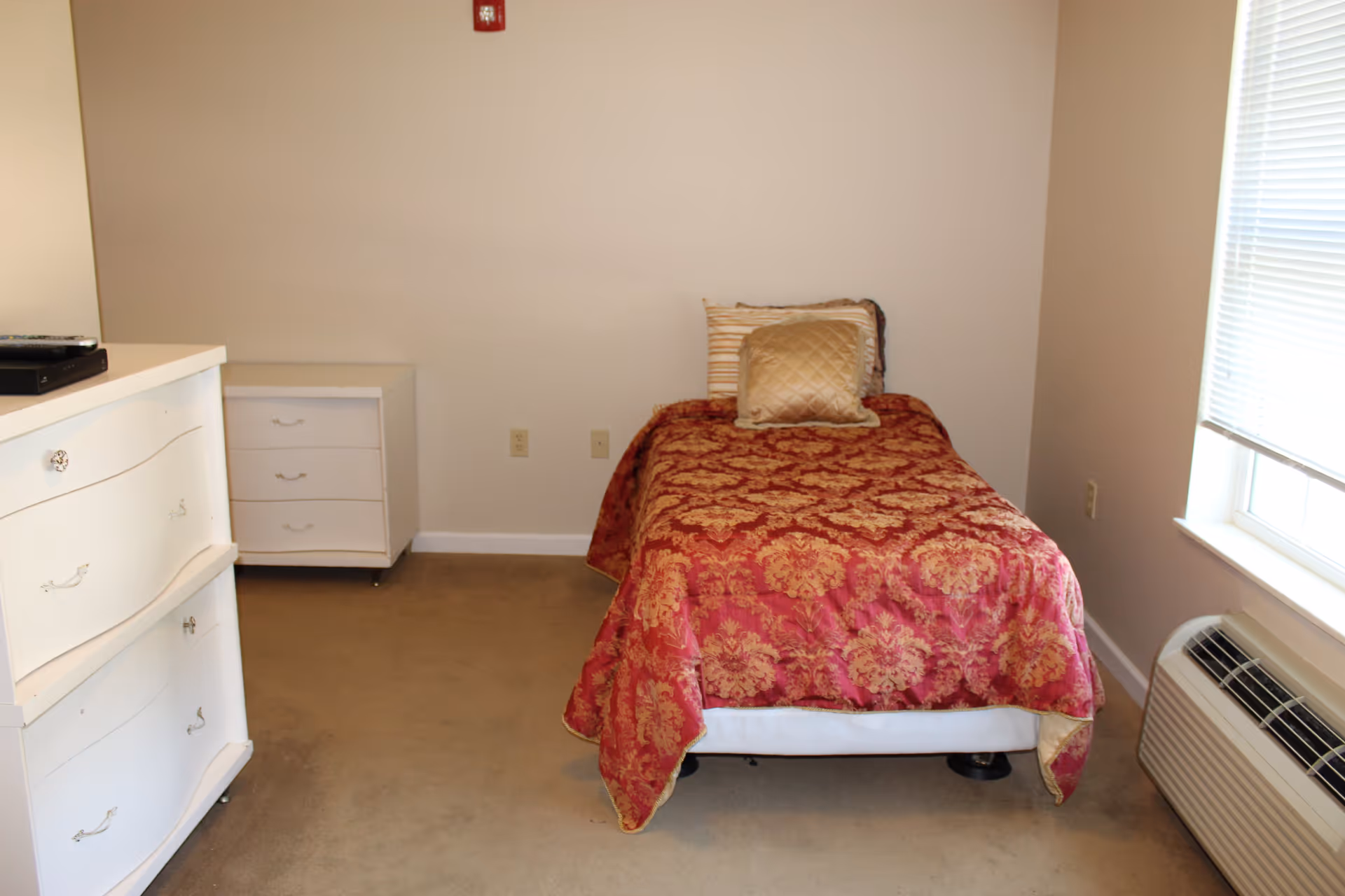 A small bedroom with a single bed covered in a red and gold patterned bedspread and two pillows. There is a white dresser with drawers on the left side and a small white nightstand with three drawers against the back wall. A window with blinds is on the right side, and below it is a wall-mounted air conditioning unit. The walls are plain and light-colored, and the floor is carpeted.