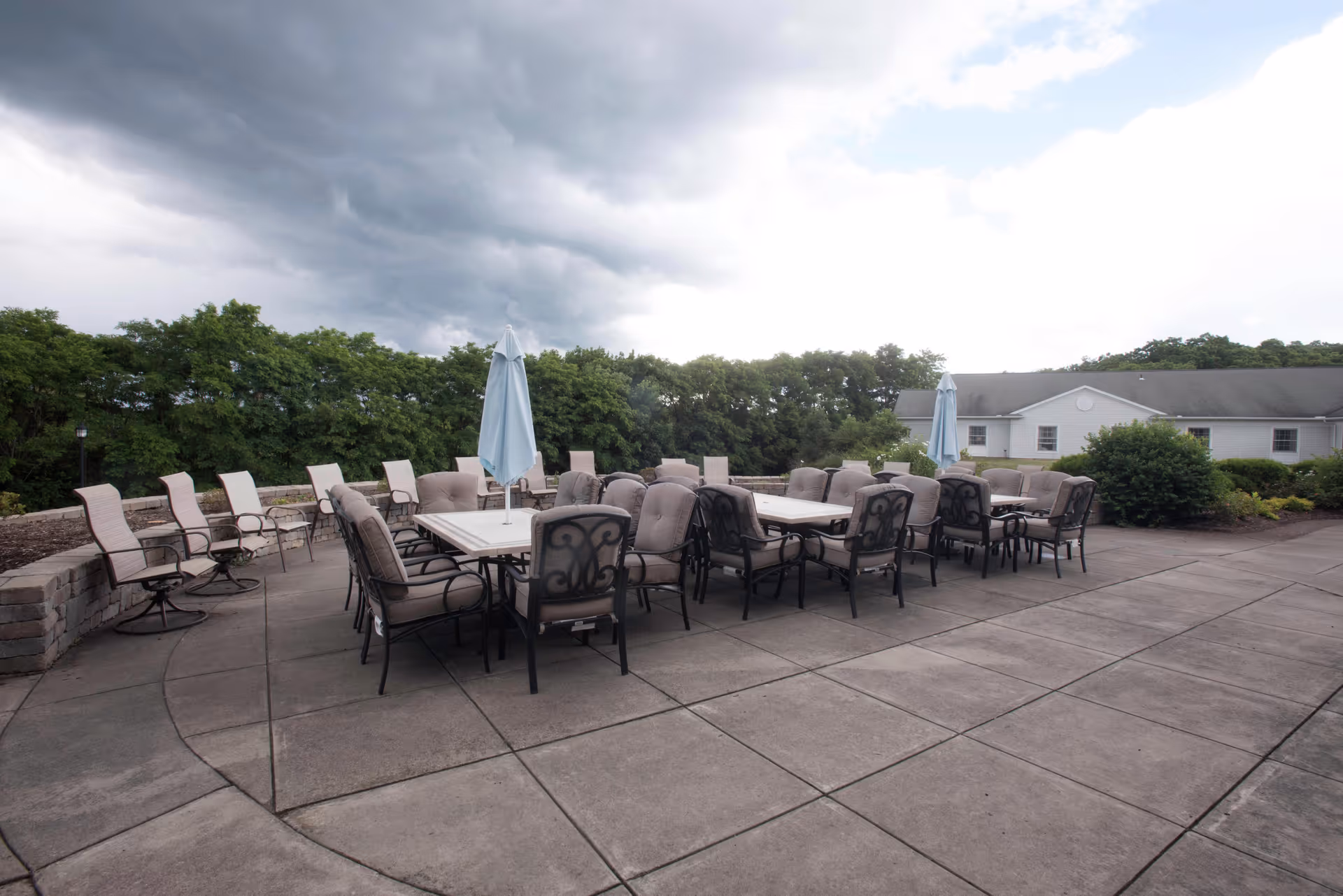 Outdoor patio area with multiple cushioned chairs and tables, some with closed umbrellas, surrounded by greenery and a building in the background under a cloudy sky.