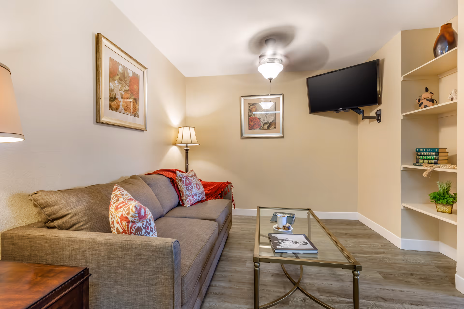 A cozy living room with a beige sofa adorned with patterned pillows and a red throw blanket. There is a glass coffee table in front of the sofa with a book, a cup, and a plate with a pastry. The room has two framed floral paintings on the walls, a floor lamp beside the sofa, a ceiling fan with a light, a wall-mounted flat-screen TV, and built-in shelves with decorative items and books.
