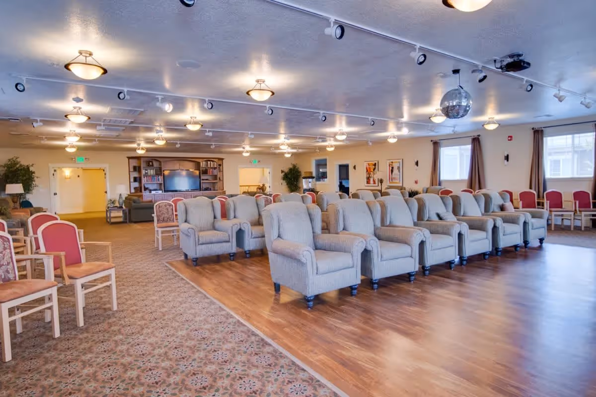 A large, well-lit common area with rows of beige upholstered armchairs arranged on a wooden floor section, surrounded by additional chairs with red cushions on a carpeted floor. The room features ceiling lights, a disco ball, windows with curtains, and a bookshelf with a TV in the background.