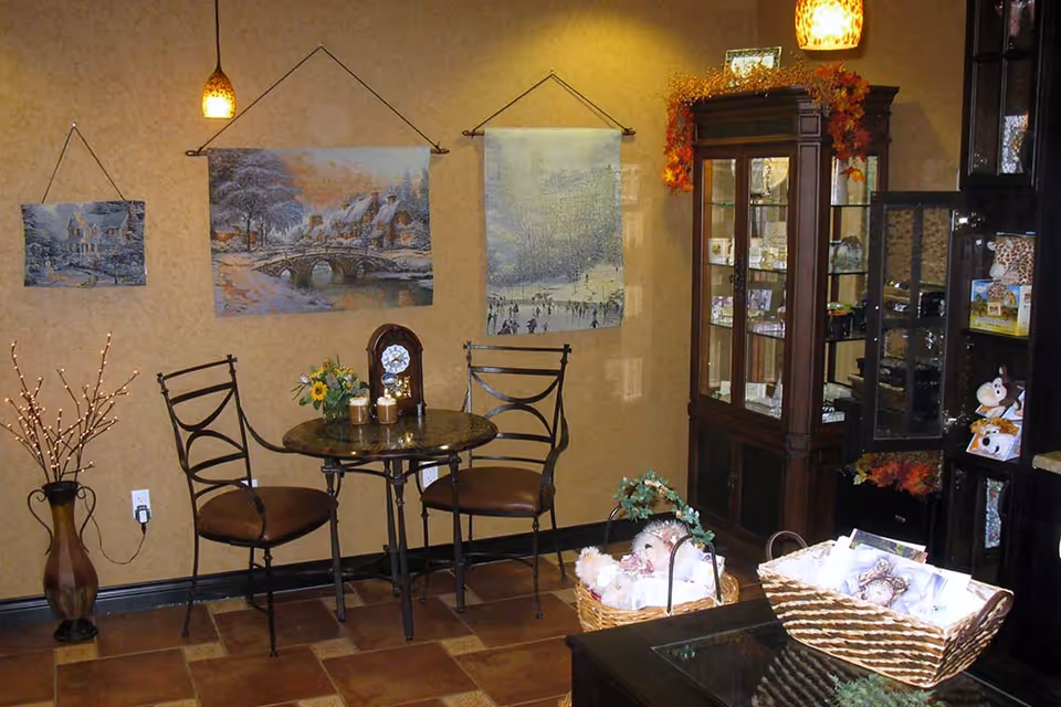 A cozy interior corner with a small round table and two metal chairs with brown cushions. The wall behind features three hanging tapestries depicting winter scenes. A wooden cabinet with glass doors displays various items and is decorated with autumn leaves. A vase with decorative branches stands on the floor, and baskets with plush toys and other items are placed on a dark wooden surface in the foreground.