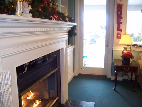 Cozy living room with a lit fireplace, holiday decorations on the mantel, a side table with a lamp and a glass door leading outside.