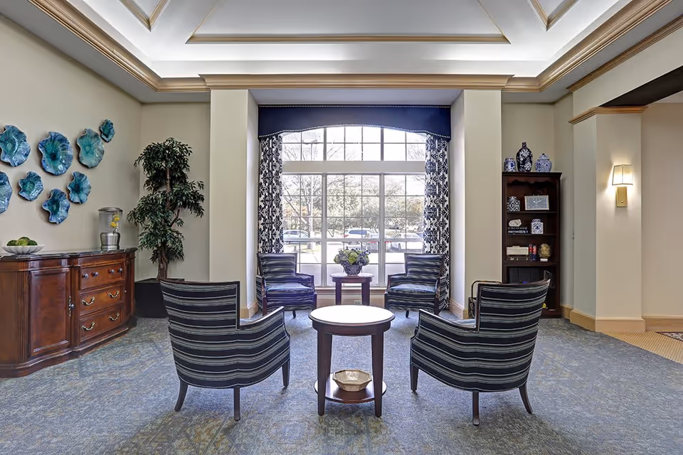 A cozy seating area in a senior living facility with four striped armchairs arranged around two small round wooden tables. Behind the chairs is a large window with patterned curtains and a blue valance, letting in natural light. To the left, there is a wooden sideboard with decorative items and a potted plant. On the right, a wooden bookshelf holds various decorative vases and books. The room has a carpeted floor and soft wall lighting.
