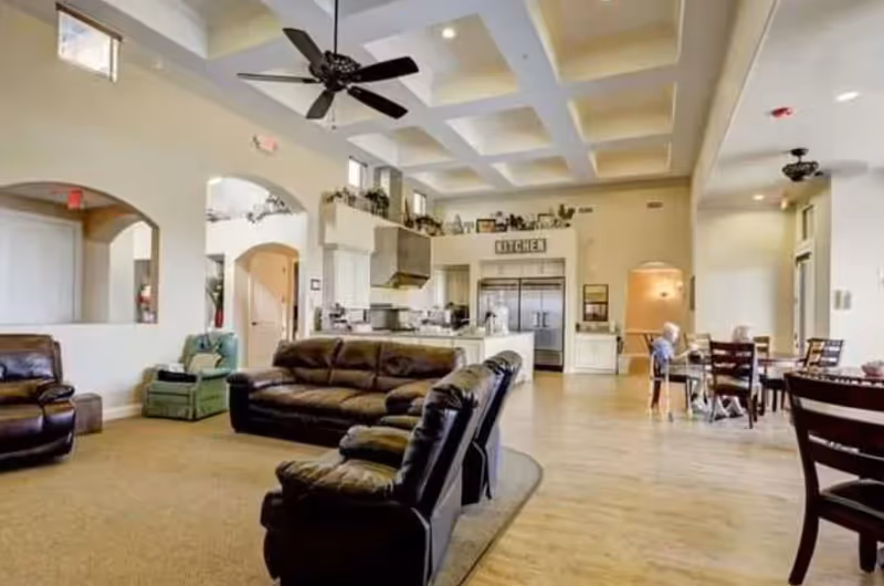 Spacious communal living area with leather sofas, a high coffered ceiling, ceiling fan, and an open kitchen and dining area in the background.