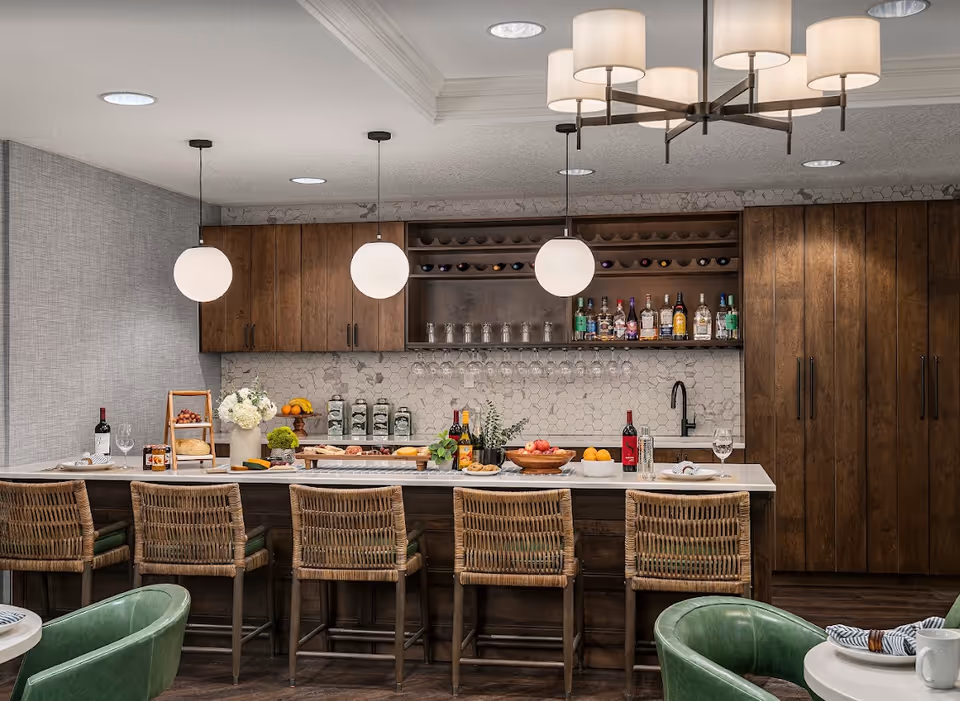 A modern kitchen area with a large island featuring six wicker bar stools. The island countertop is decorated with bottles of wine, fruit bowls, flowers, and plates. Behind the island, there are wooden cabinets, a white hexagonal tile backsplash, hanging wine glasses, and shelves stocked with liquor bottles and wine bottles. Three round pendant lights hang above the island, and a chandelier with multiple lampshades is visible on the ceiling. Two green chairs and a small table are partially visible in the foreground.