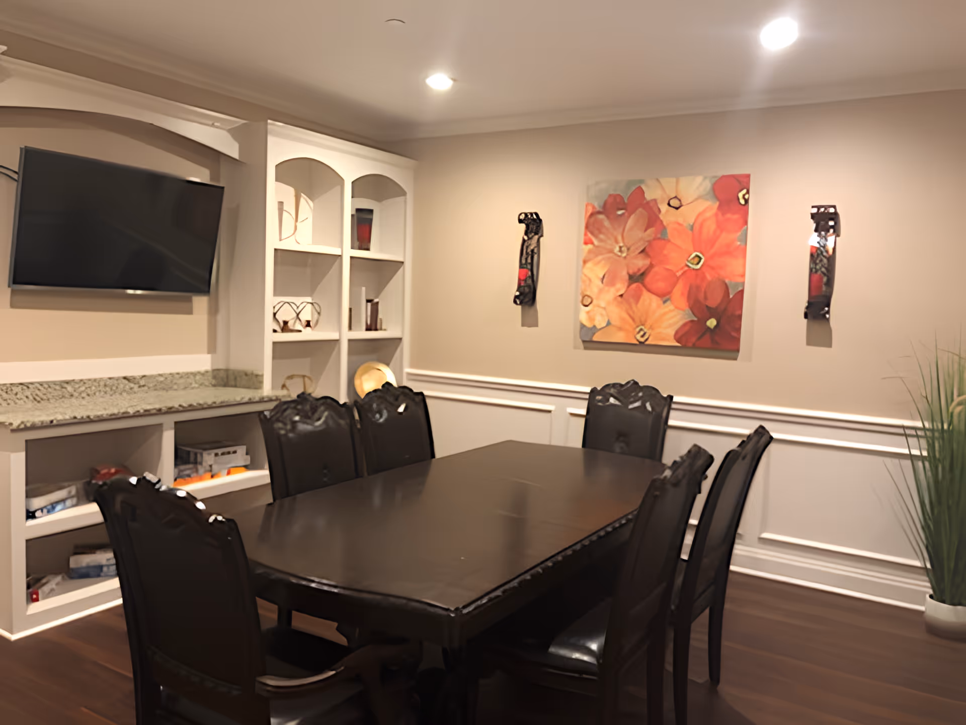 Dining room with a dark wood table and chairs, a wall-mounted TV, built-in shelving, and floral artwork on beige walls.