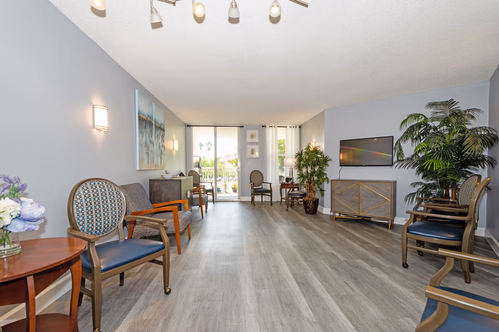 Spacious senior living lounge with arranged chairs, side tables, plants, a wall-mounted TV, and sliding glass doors to a patio.