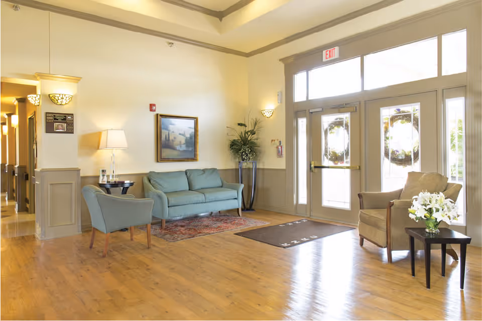 A bright and welcoming nursing facility lobby with wooden floors, a green sofa, a green armchair, a beige armchair, a small table with a vase of white flowers, a framed landscape painting on the wall, and glass double doors with exit signs above them.