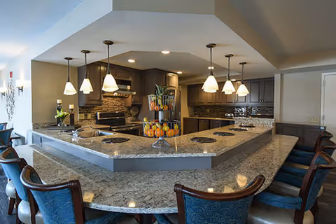 Modern kitchen area with a large granite countertop island surrounded by blue cushioned chairs. The kitchen features dark wood cabinets, stainless steel appliances, pendant lighting, and a decorative centerpiece with pumpkins on the island.