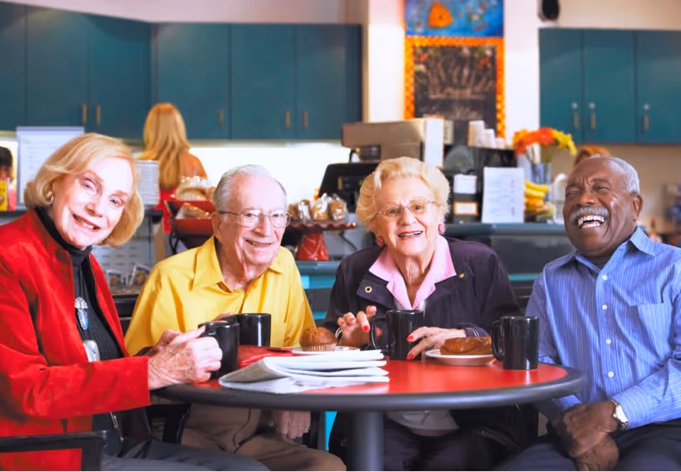 Four older adults sit around a red table in a common dining area, smiling with coffee mugs and pastries.