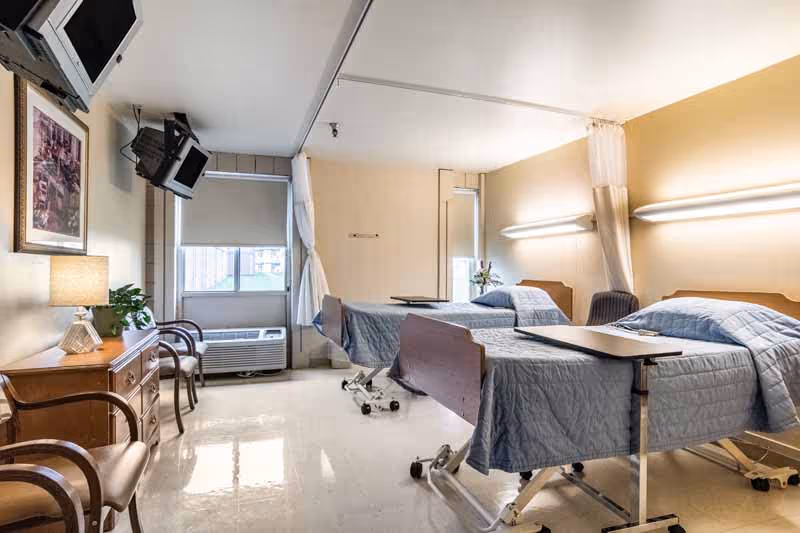 A bright senior care room featuring two hospital-style beds with blue bedspreads, chairs, a dresser, and wall-mounted TVs.