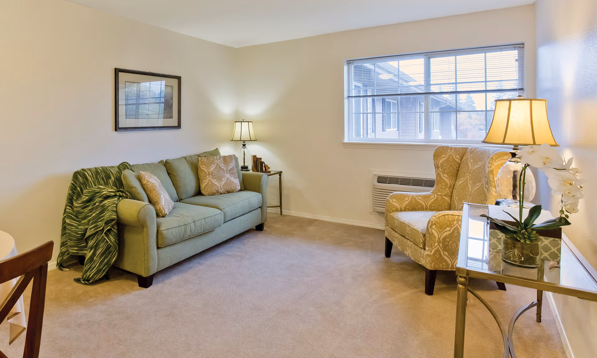 A cozy living room with a green sofa adorned with patterned cushions and a green throw blanket. Next to the sofa is a small side table with a lamp and books. Across from the sofa is a beige patterned armchair beside a glass-top side table with a lamp and a white orchid plant. A large window with blinds lets in natural light, and the room has beige carpet and light-colored walls.