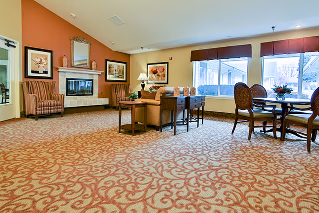 A spacious living room area with patterned carpet, a fireplace with a mirror and framed artwork above it, a striped armchair, a sofa with a side table and lamp, and a round dining table with four chairs near large windows with red valances.