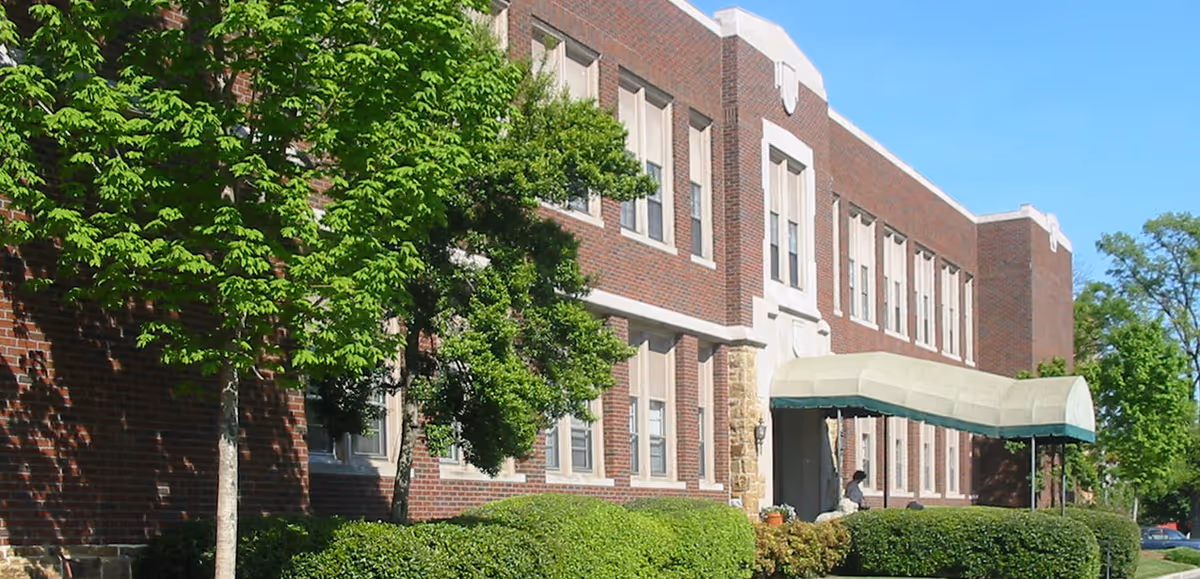 Exterior view of a two-story brick building with multiple windows, surrounded by green trees and bushes under a clear blue sky. A covered entrance canopy extends from the building.