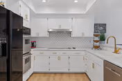 Modern kitchen with white cabinets, gold handles, a black refrigerator, built-in oven, microwave, white countertop with a gold faucet, and a coffee maker on the counter. The backsplash features a white geometric tile pattern, and the floor is light wood.
