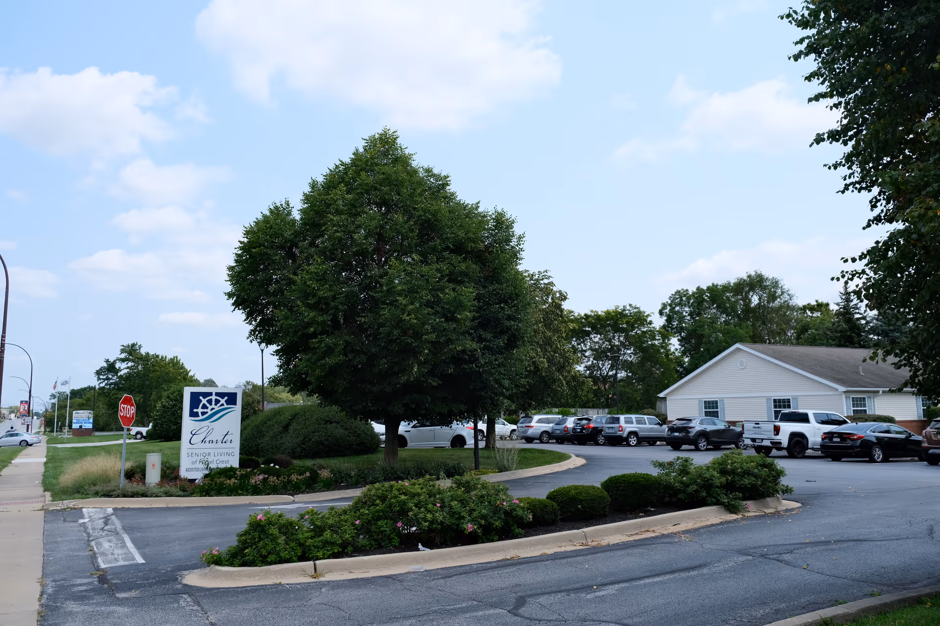 Parking lot area of Charter Senior Living of Hazel Crest with several parked cars, a large tree, bushes, and a sign displaying the facility's name. A stop sign and sidewalk are visible on the left side under a partly cloudy sky.