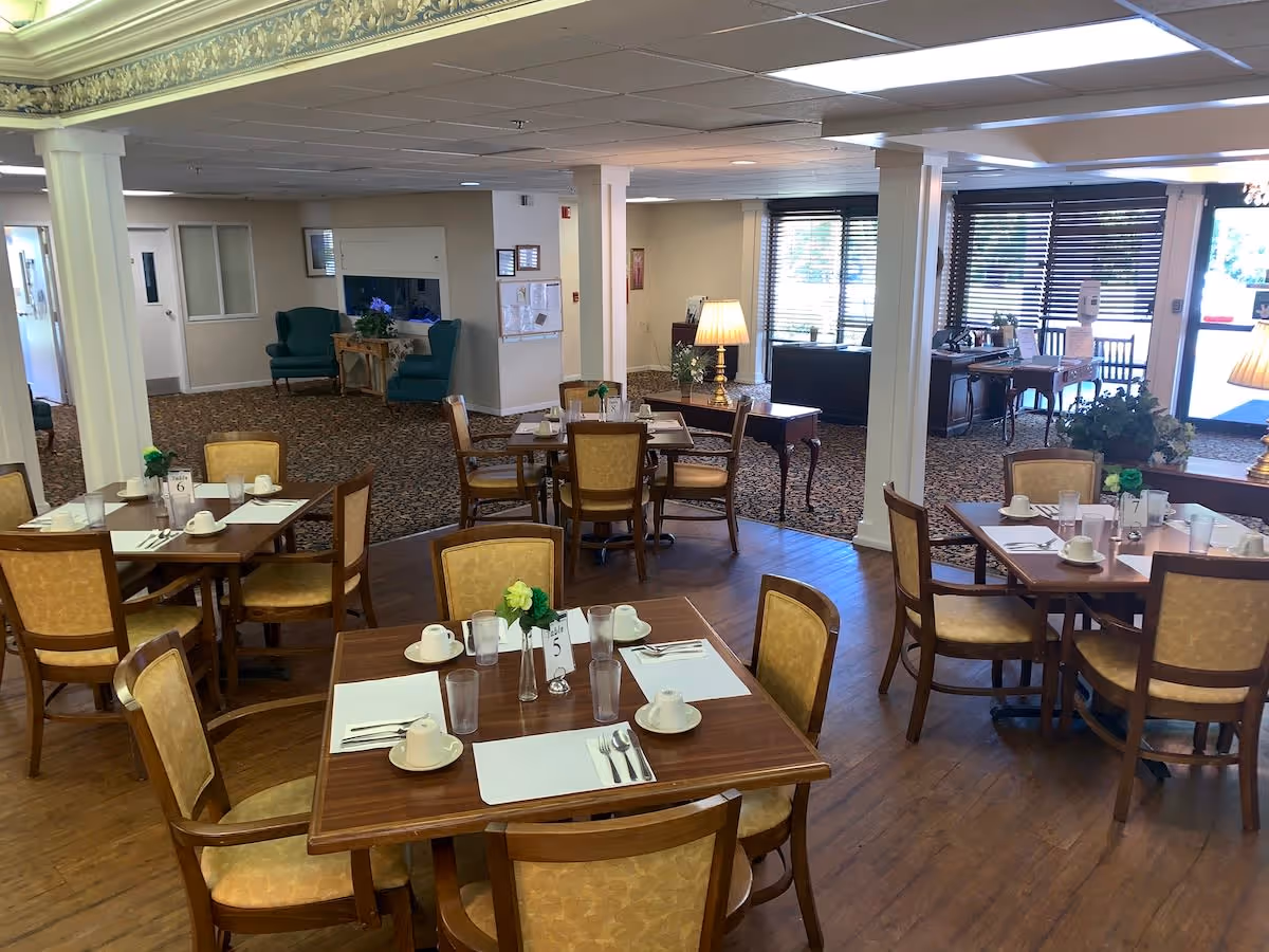 Dining area in Oakview Terrace Assisted Living and Memory Care with several wooden tables and chairs arranged neatly. Each table is set with placemats, cups, glasses, and utensils. The room has wooden flooring and carpeted sections, with large windows covered by blinds allowing natural light. There are also a few armchairs and side tables with lamps in the background.