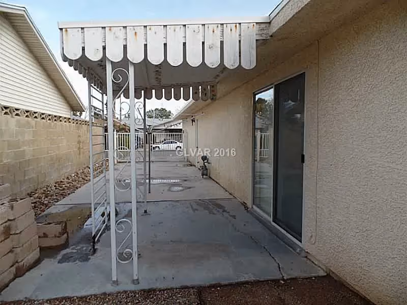 Covered concrete patio alongside a stucco house with a scalloped metal awning, decorative metal posts, and a sliding glass door.