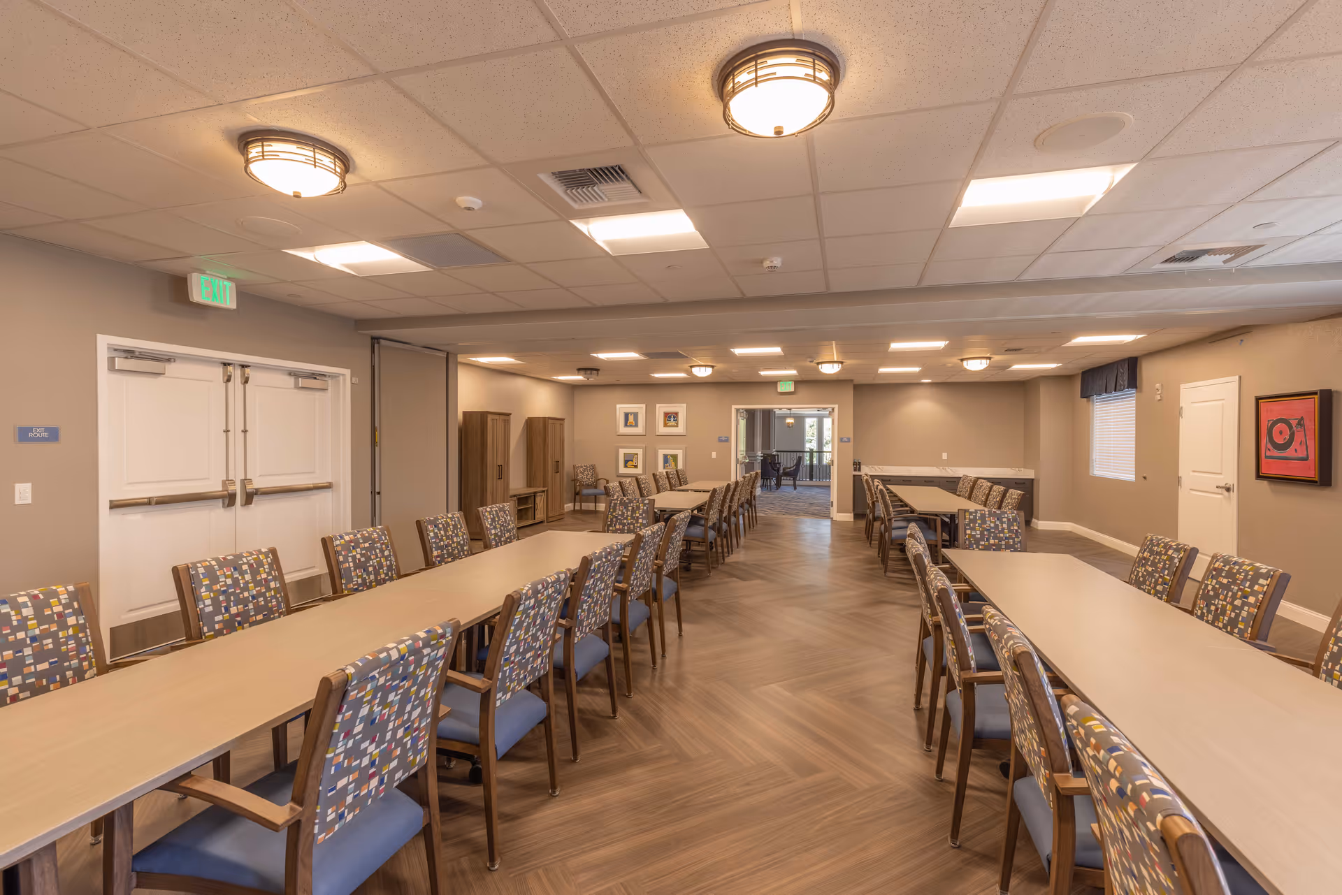 Spacious community dining room with long rectangular tables and patterned chairs under ceiling lights.