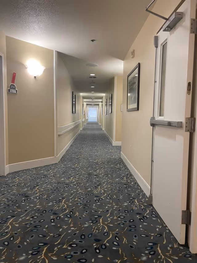 Long indoor hallway in a senior living facility with patterned blue carpet, beige walls, handrails along the walls, framed pictures, and ceiling lights. A white door is open on the right side near the foreground.