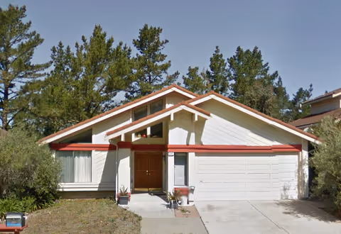 Front exterior view of a single-story house with a white facade and red trim. The house has a double garage door on the right, a central entrance with double wooden doors, and large windows on the left. Tall pine trees are visible in the background under a clear blue sky.