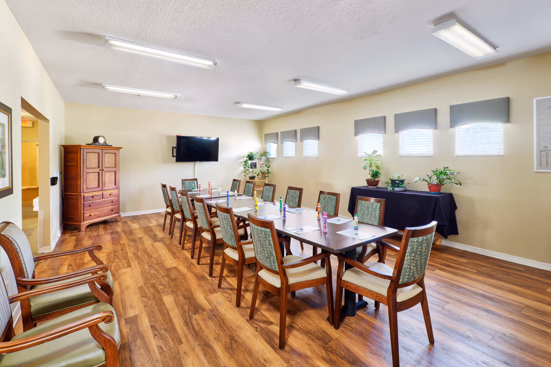 A well-lit meeting or activity room with a long rectangular table surrounded by wooden chairs with cushioned seats. The table has papers and colorful figurines placed on it. The room has wooden flooring, beige walls, and several small windows with gray valances. There is a flat-screen TV mounted on the wall, a wooden cabinet, some potted plants on a table covered with a black cloth, and two armchairs with wooden frames and green cushions near the entrance.