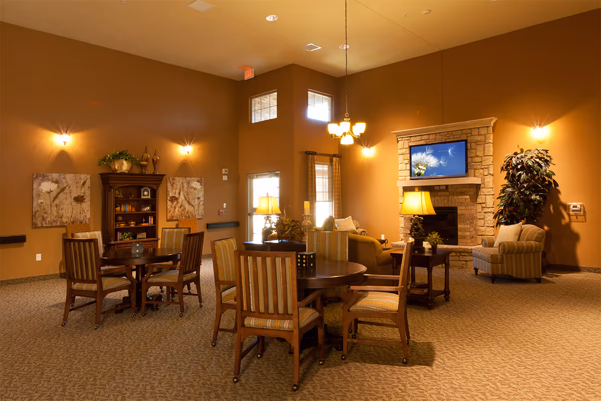 Warmly lit common lounge with round tables and chairs, upholstered seating, a stone fireplace with a TV, and decorative accents.