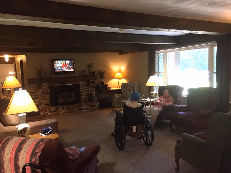 A cozy living room with wooden ceiling beams, a brick fireplace, and several armchairs. Two elderly individuals are seated, one in a wheelchair and the other in an armchair near a large window letting in natural light. Multiple lamps provide warm lighting throughout the room.