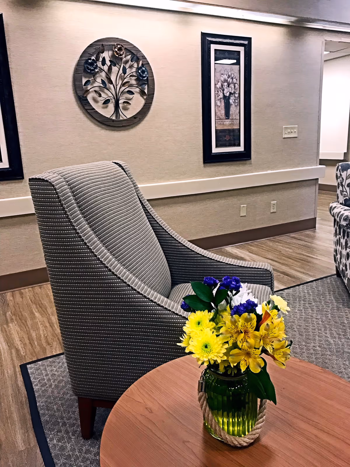 A cozy seating area in a senior living facility featuring a striped upholstered armchair next to a round wooden table with a green glass vase holding a colorful bouquet of yellow, purple, and white flowers. The background shows beige walls adorned with framed floral artwork and a decorative metal wall hanging, with wood-look flooring and a patterned rug.