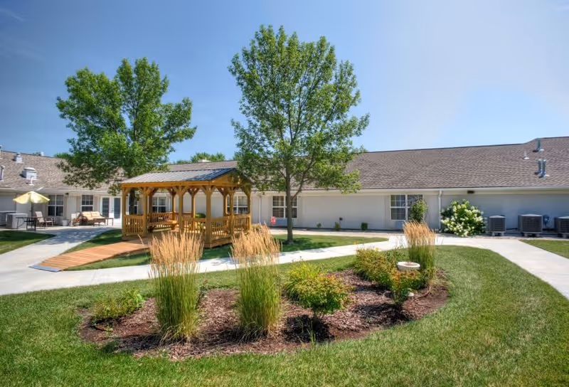 Outdoor courtyard area at Vintage Park at Hiawatha featuring a wooden gazebo, green trees, landscaped garden beds with ornamental grasses and shrubs, and a paved walkway surrounding the space. The building exterior with windows and air conditioning units is visible in the background under a clear blue sky.