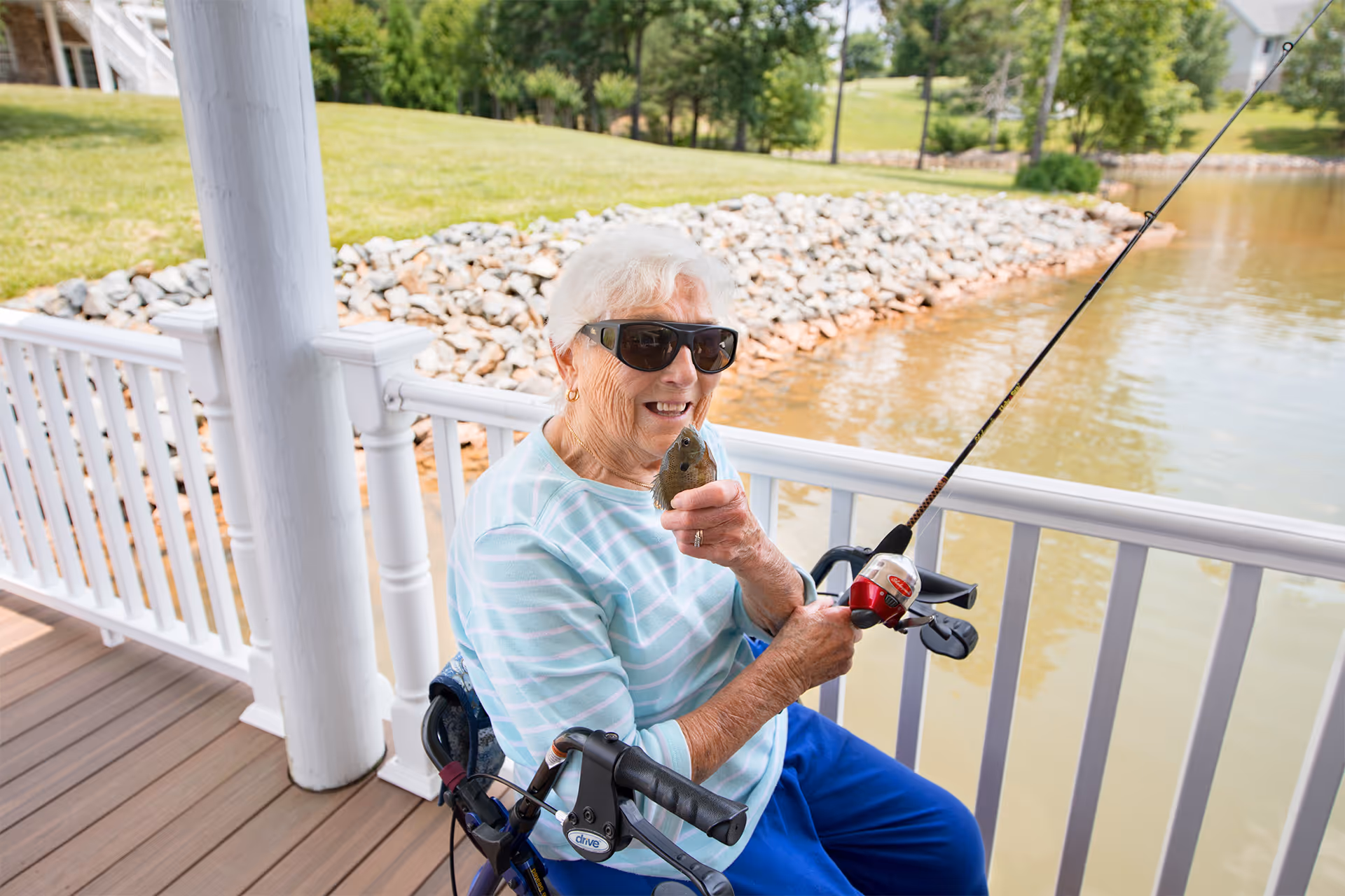 An elderly woman wearing sunglasses and a light blue striped shirt is sitting on a porch by a lake, holding a fishing rod in one hand and a small fish in the other. She is smiling and appears to be enjoying fishing. The porch has white railings and wooden flooring, with a grassy area and rocks visible in the background.