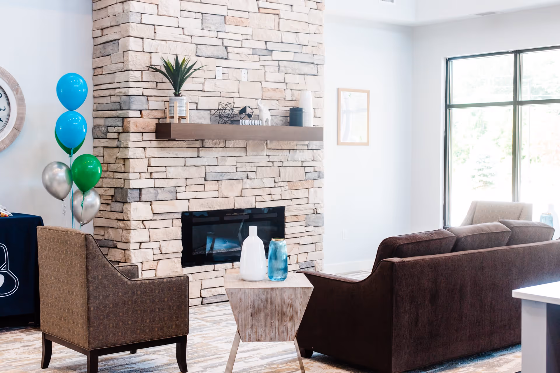 A cozy living room area with a stone fireplace, a wooden mantel decorated with small plants and ornaments, a brown upholstered sofa, a patterned armchair, a small wooden side table with decorative vases, and a large window letting in natural light. There are also blue, green, and silver balloons near the armchair.
