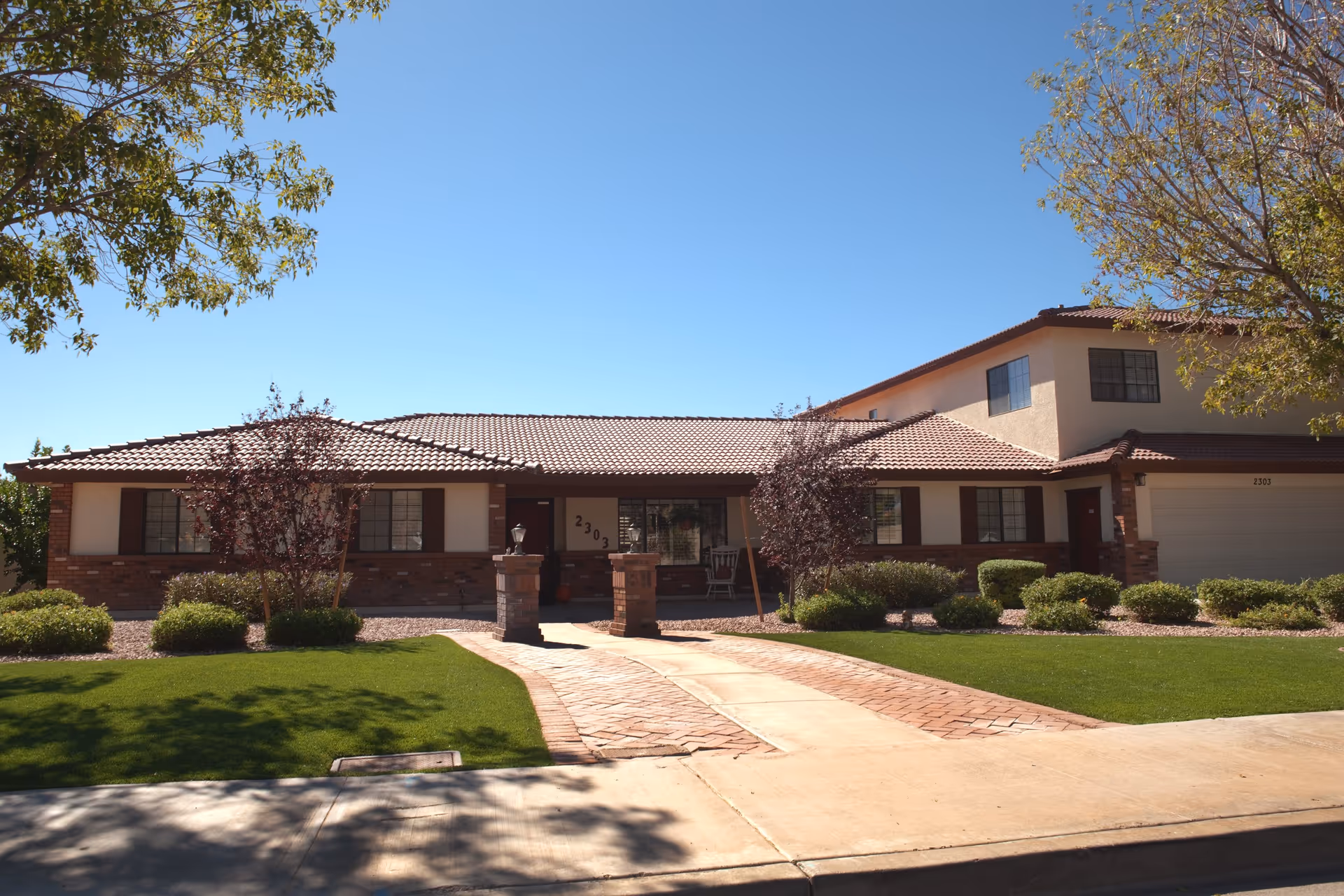 Front exterior view of a single-story residential building with a tiled roof, brick and stucco walls, and several windows. The house number 2303 is visible near the entrance. There is a paved walkway leading to the front door, flanked by two brick pillars with lanterns. The lawn is well-maintained with green grass, bushes, and small trees. The sky is clear and blue.