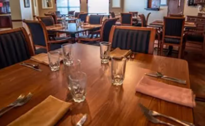 Wooden dining tables set with glasses, napkins, and silverware in a dining room with upholstered chairs.