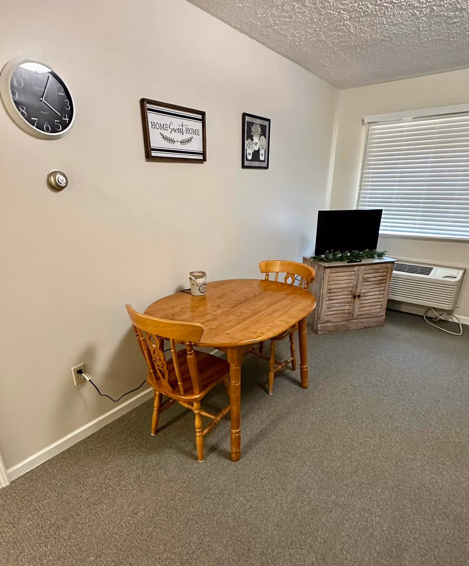 A small dining area with a wooden oval table and two wooden chairs. On the table is a small decorative item. The wall behind the table has a round clock, a thermostat, and two framed pictures, one with the text 'Home Sweet Home' and another with a plant illustration. To the right, there is a wooden cabinet with a flat-screen TV on top, a window with closed blinds, and an air conditioning unit below the window.