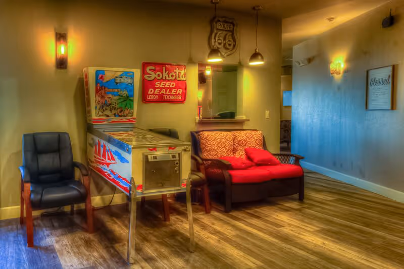 A cozy interior corner of a senior living facility featuring a vintage pinball machine with nautical designs, a black cushioned chair, and a small loveseat with red cushions. The walls are decorated with a Route 66 sign, a red Sokota Seed Dealer sign, and a framed welcome message. Warm lighting fixtures illuminate the space, and the floor is covered with wood-patterned vinyl.