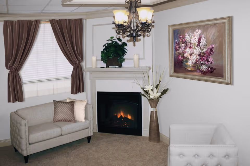 A cozy living room with a beige loveseat and armchair, a fireplace with a plant and candles on the mantel, a large floral painting on the wall, brown curtains on the window, and a chandelier hanging from the ceiling.