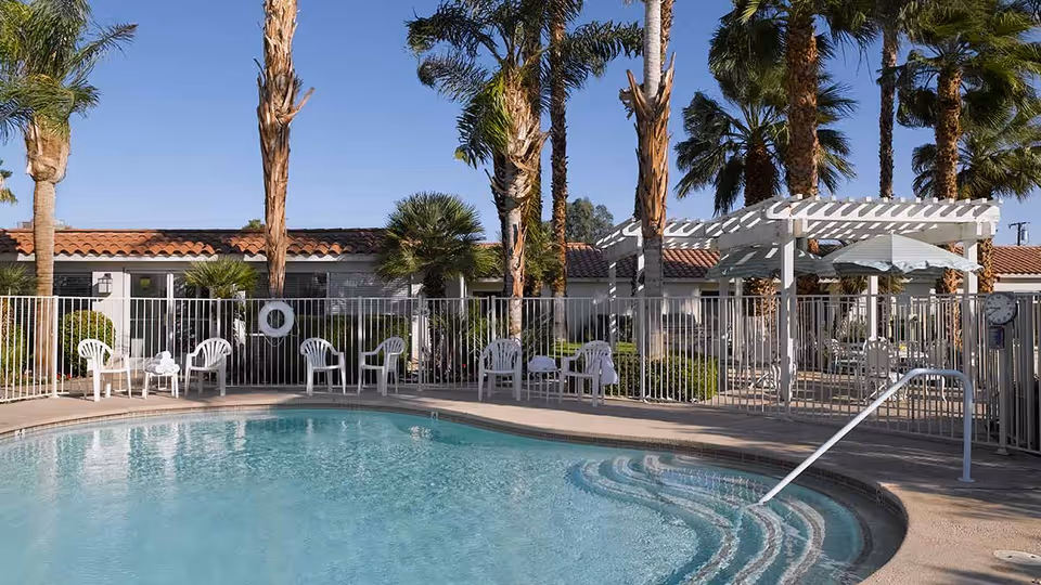 Outdoor swimming pool area with clear blue water, surrounded by white metal fencing. Several white plastic chairs and tables are placed around the pool. Tall palm trees and a white pergola with an umbrella provide shade. A building with a red-tiled roof is visible in the background under a clear blue sky.