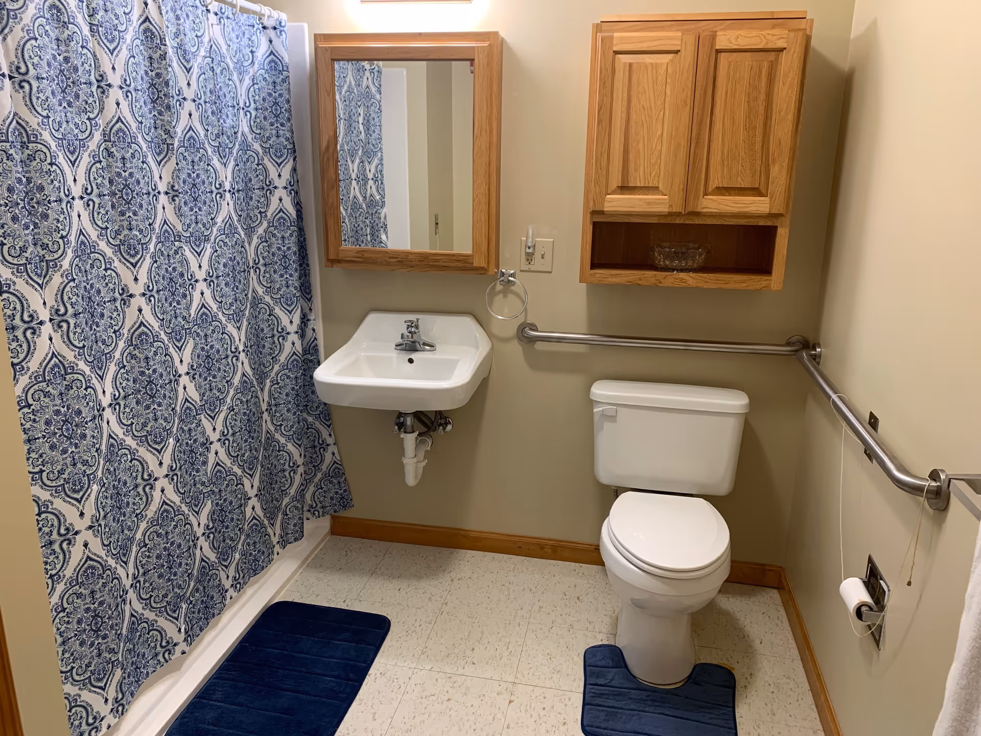 Bathroom with a toilet, wall-mounted sink, wooden medicine cabinet, grab bars, and a blue patterned shower curtain.
