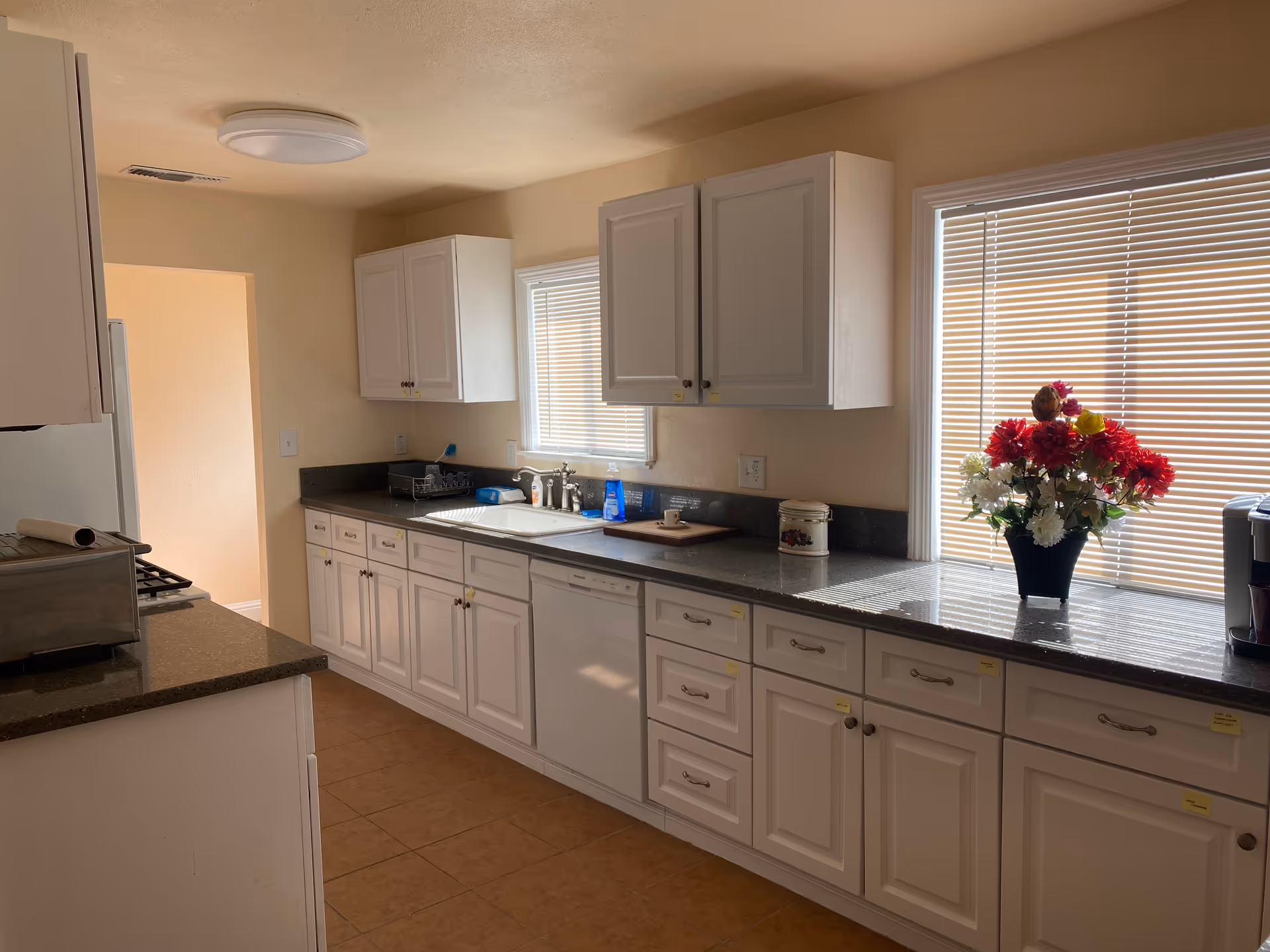 A bright kitchen with white cabinets and drawers, a gray countertop, and a tiled floor. There are two windows with blinds letting in natural light. On the countertop, there is a vase with red, white, and yellow flowers, a coffee maker, a cutting board, and various kitchen items near the sink. The kitchen has a clean and organized appearance.