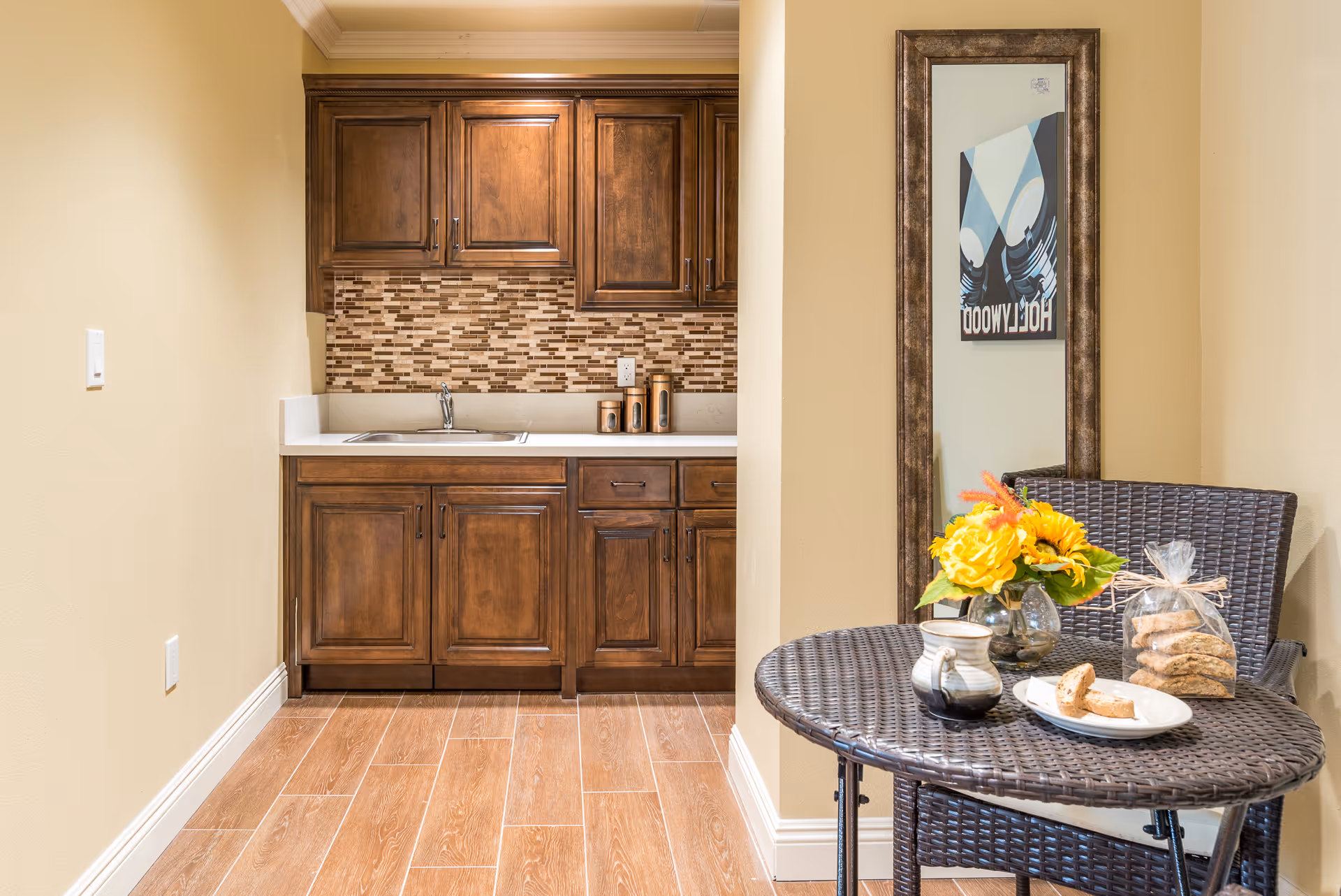 A small kitchenette with wooden cabinets, a sink, and a mosaic tile backsplash. To the right, there is a round wicker table with a matching chair, a vase with yellow flowers, a ceramic mug, and a plate with biscotti. A tall framed mirror is mounted on the wall behind the table, reflecting a partial view of a Hollywood-themed poster.