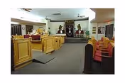 Interior room with wooden pews and seating facing a raised platform with decorative elements at the far end.