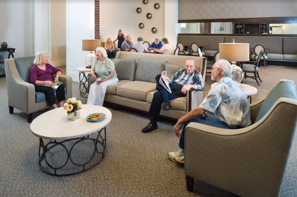 Elderly residents sitting and chatting in a well-furnished senior living lounge with sofas, chairs and a coffee table.